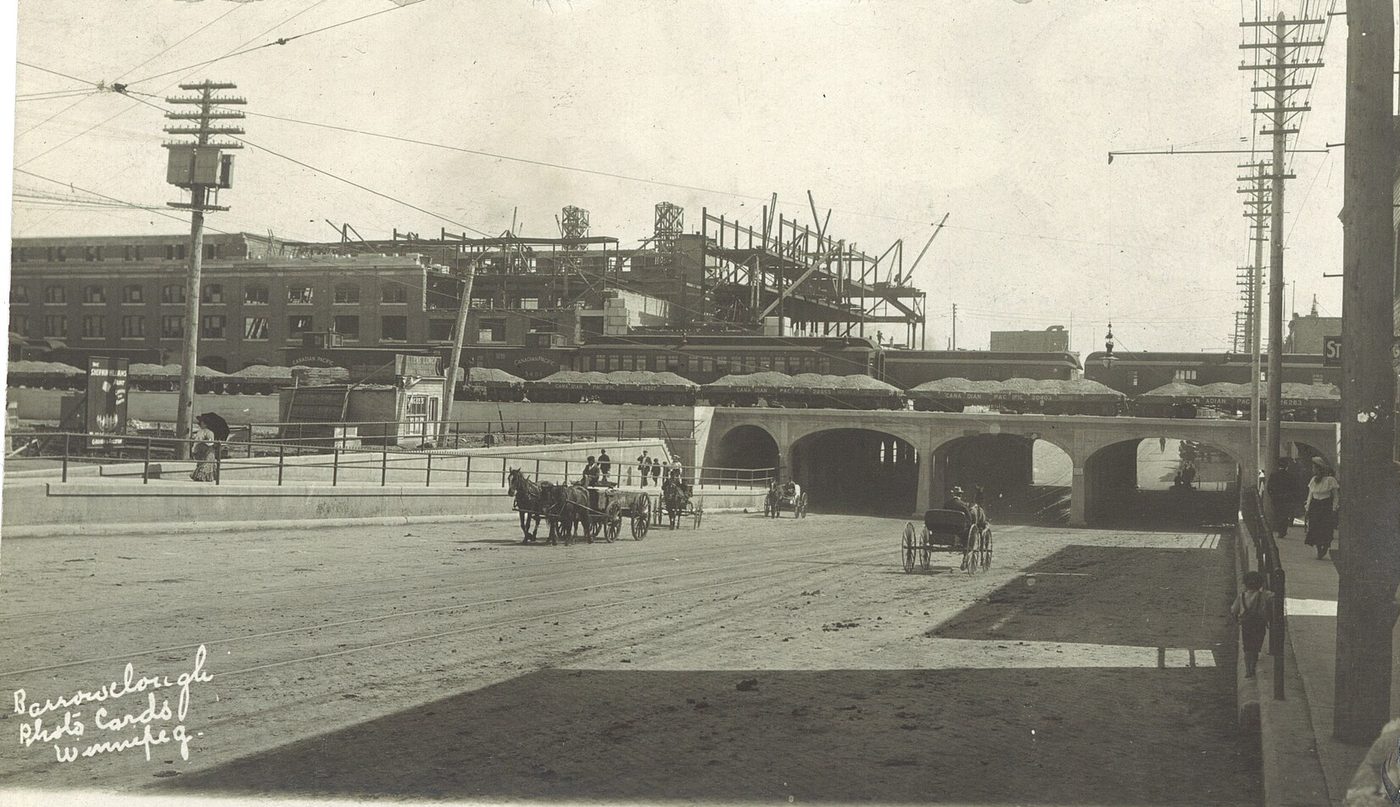 Old photograph of a wide unpaved street leading to a railway underpass, with horse-drawn wagons and a train crossing above